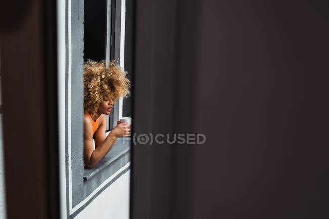 Curly ethnique femme tenant tasse et regardant par la fenêtre — Photo de stock