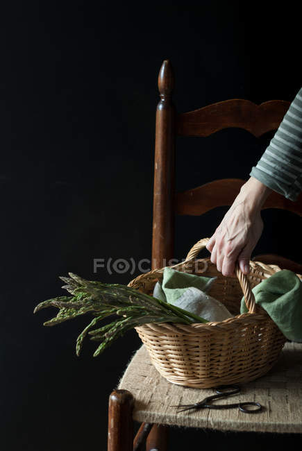 Menschliche Hand hält Korb mit frischem grünen Spargel auf Stuhl auf schwarzem Hintergrund — Stockfoto