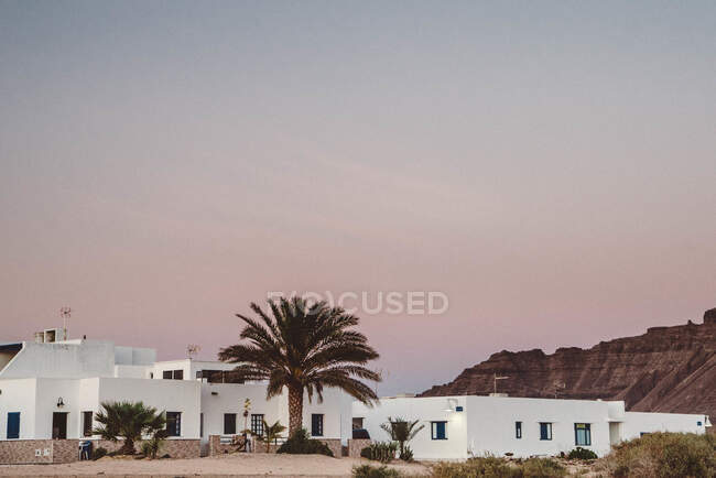 Vista panoramica di un piccolo edificio bianco circondato da palme situato nella tropicale La Graciosa, Isole Canarie al tramonto con montagne e sorprendente cielo pastello sullo sfondo — Foto stock