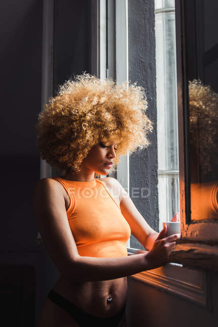 Молодая чувственная женщина в нижнем белье с афроволосами, стоящая у окна с чашкой — стоковое фото
