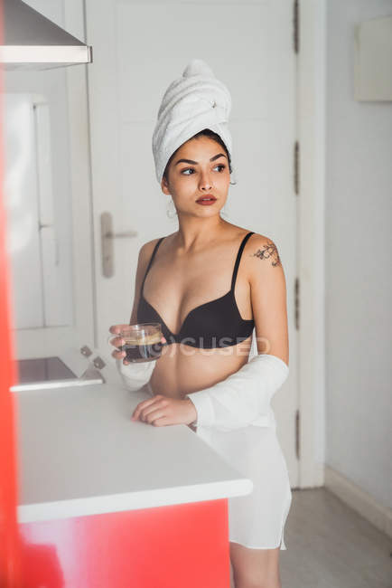 Dreamy young woman in black bra and towel on hair holding cup of coffee in kitchen — Stock Photo