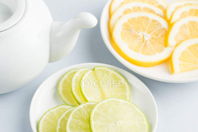 Scheiben reife saftige Limette und Zitrone auf Untertassen auf weißem Hintergrund mit Teekanne — Stockfoto