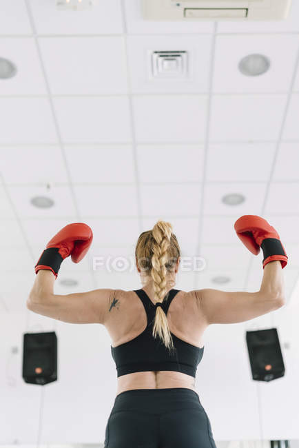 Назад вид м'язистої жінки в червоних боксерських рукавичках, що показують біцепси, стоячи на розмитому тлі тренажерного залу — стокове фото