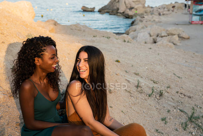 Smiling multiethnic women embracing and chilling on coastline in sunlight — Stock Photo
