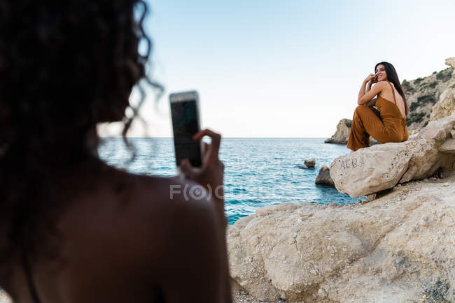 Mulher negra tirando foto com smartphone de amigo elegante sentado no penhasco rochoso do litoral no verão — Fotografia de Stock