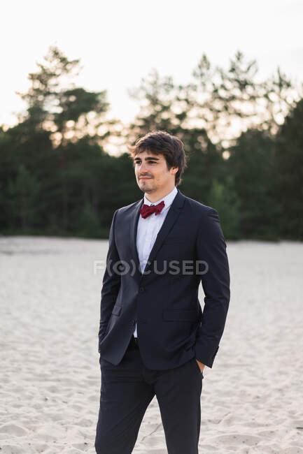 Hombre adulto con elegante traje negro con pajarita y de pie en la playa con las manos en los bolsillos mirando a la cámara - foto de stock