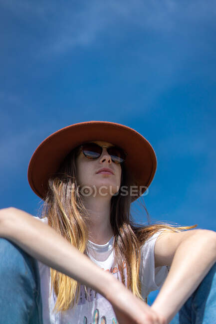 Da sotto colpo di giovane donna sicura di sé con i capelli lunghi indossa abbigliamento casual con occhiali da sole e cappello seduto sotto il cielo blu — Foto stock