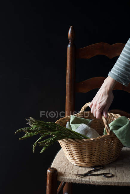 Mão humana segurando cesta de espargos verdes frescos na cadeira no fundo preto — Fotografia de Stock