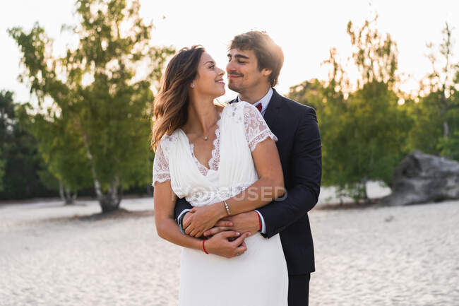 Aimant l'homme embrassant belle mariée dans une robe élégante et se regardant tout en se tenant debout sur la côte sablonneuse au soleil — Photo de stock