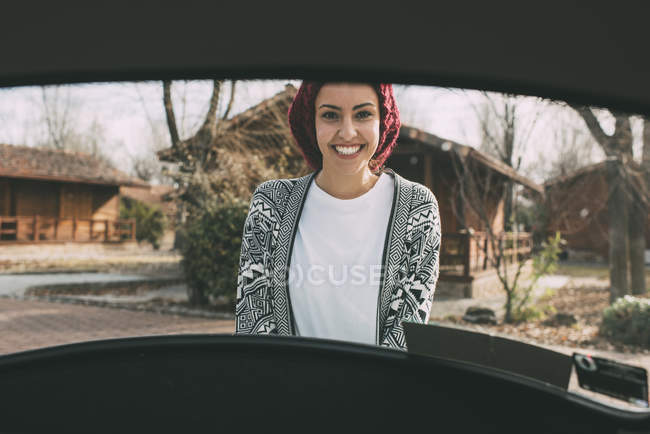 Junge Frau mit Wollmütze steht im Grünen hinter Auto — Stockfoto