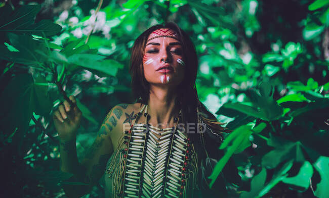 Jolie jeune femme avec des peintures indiennes traditionnelles sur le visage regardant la caméra et debout dans la forêt — Photo de stock