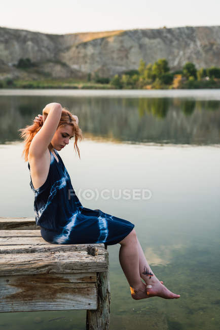 Sensuale giovane donna in abito seduto sul molo al lago — Foto stock