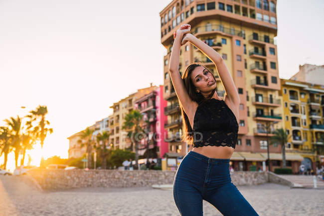 Young brunette woman in jeans and black top posing on beach at sunset — Stock Photo