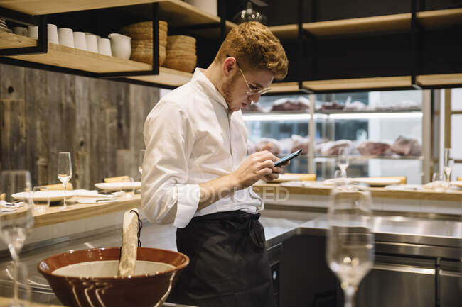 Side view of young man in cook uniform leaning on kitchen counter and using modern smartphone while standing in restaurant kitchen — Stock Photo