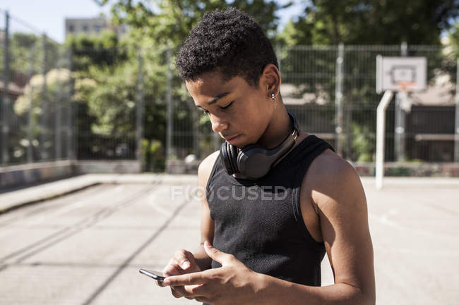 Niño afro escuchando música con smartphone y auriculares en cancha de baloncesto - foto de stock