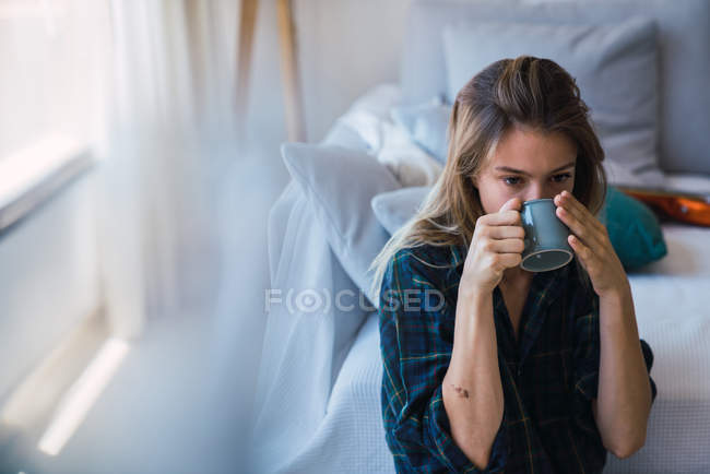 Mujer joven disfrutando de una bebida caliente mientras está sentada cerca del sofá en una habitación acogedora - foto de stock