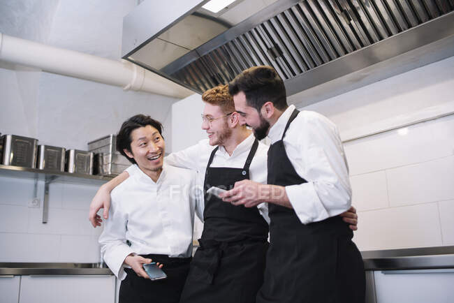 Tres hombres diversos en uniforme de cocinero riendo y navegando teléfonos inteligentes mientras están de pie en la cocina del restaurante juntos - foto de stock