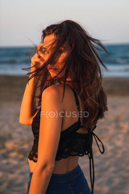 Splendida donna bruna in piedi sulla spiaggia al tramonto e guardando oltre le spalle — Foto stock