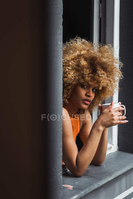 Riccio donna etnica in possesso di tazza e guardando fuori dalla finestra — Foto stock