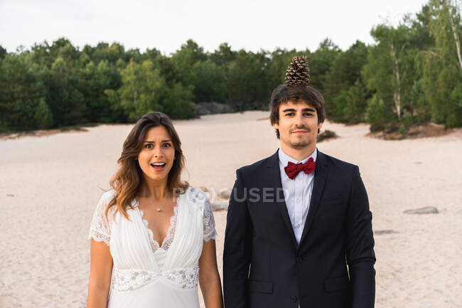 Смішний наречений з сосновим конусом на голові і виразною здивованою нареченою, що стоїть разом на узбережжі, дивлячись на камеру — стокове фото
