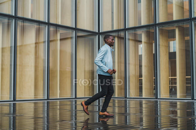 Élégant homme noir qui court sur un trottoir humide contre un bâtiment en verre — Photo de stock