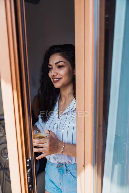 Donna bruna sorridente che tiene in mano una tazza di caffè e guarda attraverso la finestra — Foto stock