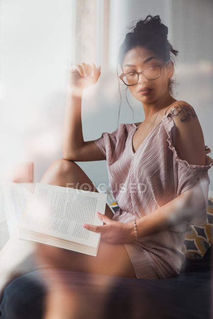 Femme brune avec livre assis sur le lit et regardant loin — Photo de stock