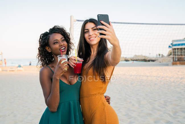 Mujeres multiétnicas de moda tomando bebidas y tomando selfie mientras con el teléfono inteligente en la playa - foto de stock