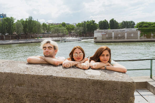 Les jeunes femmes et l'homme se détendre tout en s'appuyant sur les mains croisées sur le mur de pierre au bord de l'eau — Photo de stock