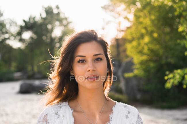 Morena encantadora vestindo vestido de noiva elegante com colar e olhando para a câmera ao ar livre — Fotografia de Stock