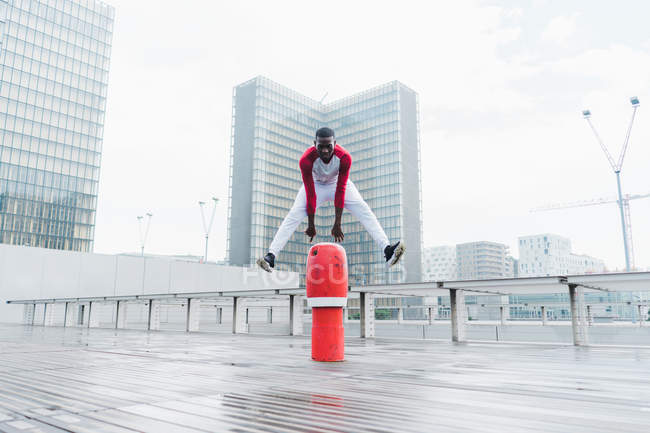 Homme ethnique fort en vêtements de sport sautant par-dessus obstacle rouge sur chaussée mouillée avec la ville moderne — Photo de stock