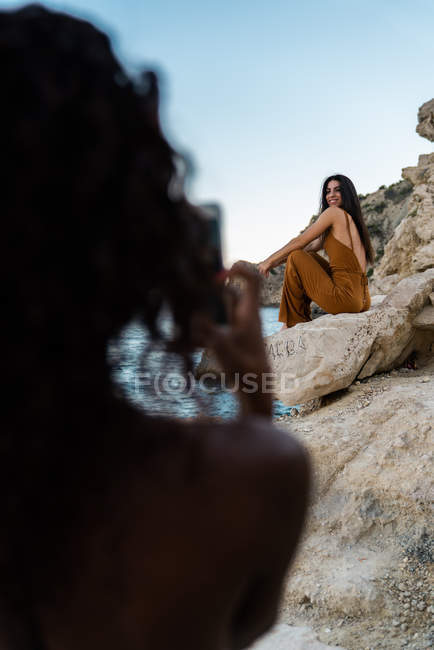 Donna nera che scatta foto con smartphone di un amico elegante seduto sulla scogliera rocciosa del mare in estate — Foto stock