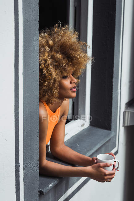 Curly mulher étnica segurando copo e olhando para fora da janela — Fotografia de Stock