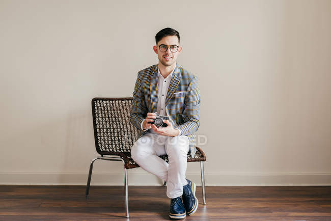 Nachdenklich stilvoller junger Mann sitzt auf Stuhl mit Retro-Kamera — Stockfoto