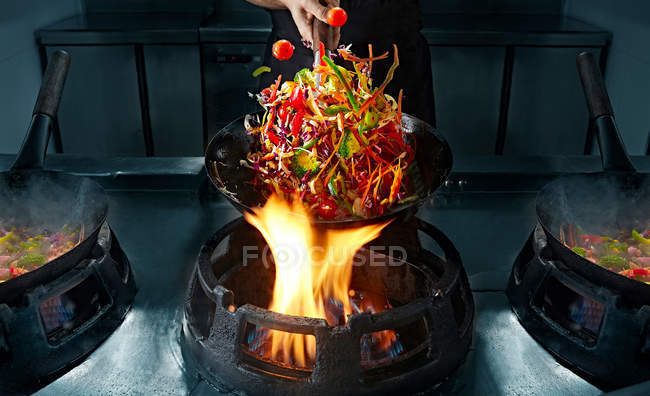 Wok de cozimento da mão da colheita na panela sobre o fogo e em movimento legumes no ar — Fotografia de Stock