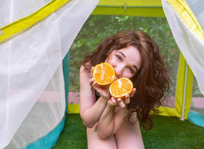 Linda mujer joven sosteniendo mitades de naranja cerca de la cara y mirando a la cámara mientras está sentado en la tienda de campaña en el jardín - foto de stock