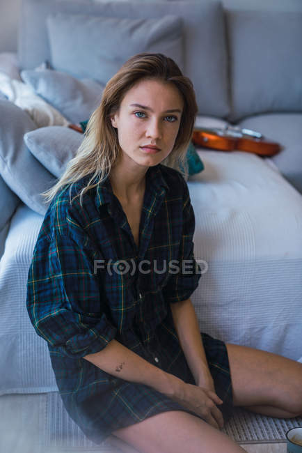 Junge Frau im karierten Oversize-Hemd sitzt auf dem Boden neben dem Sofa und blickt in die Kamera — Stockfoto