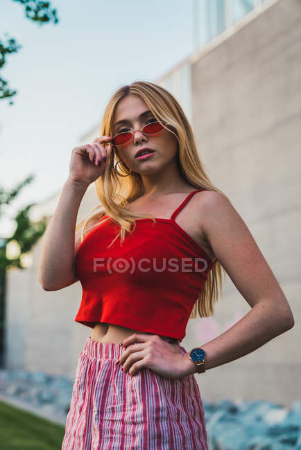 Young woman in shorts and red tank top touching sunglasses while standing on street — Stock Photo