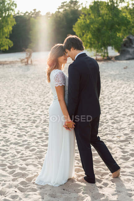 Loving man embracing beautiful bride in elegant dress and looking at each other while standing on sandy coast in sunlight — Stock Photo
