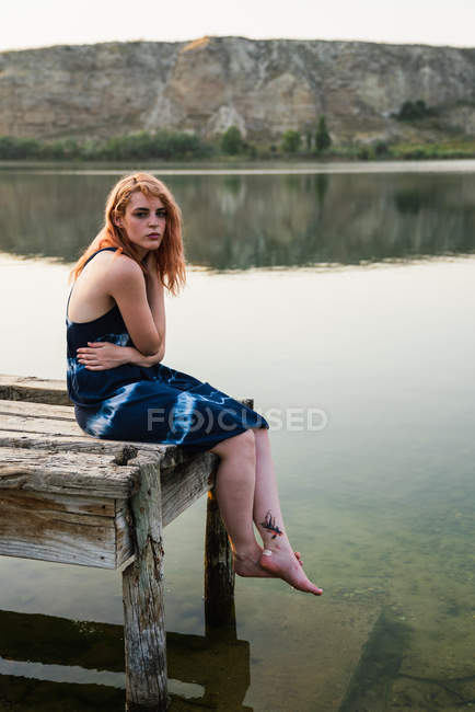 Чувственная молодая женщина в платье сидит на пирсе у озера на природе — стоковое фото