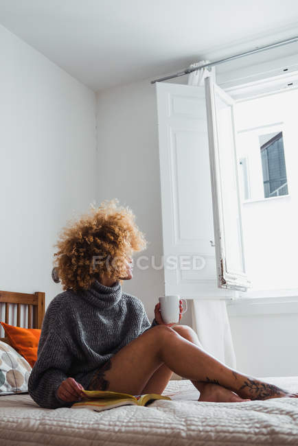Ethnic woman relaxing with coffee and book on bed — Stock Photo