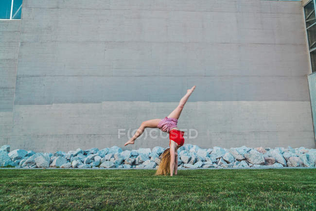 Joven mujer haciendo handstand en hierba contra pared de hormigón y piedras - foto de stock