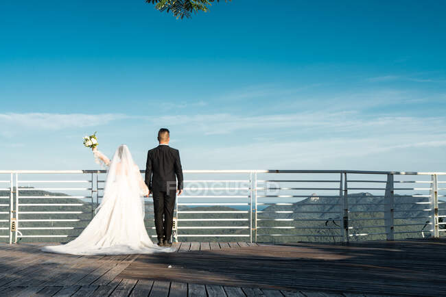 Vista trasera de la novia sosteniendo ramo y de pie con el novio en la terraza del mirador contra el paisaje, Phoenix Park, China - foto de stock