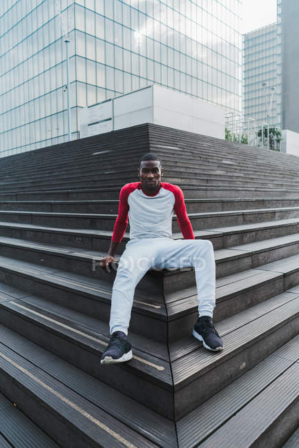 Young ethnic man in sportswear sitting on stairs and against glass buildings in city — Stock Photo