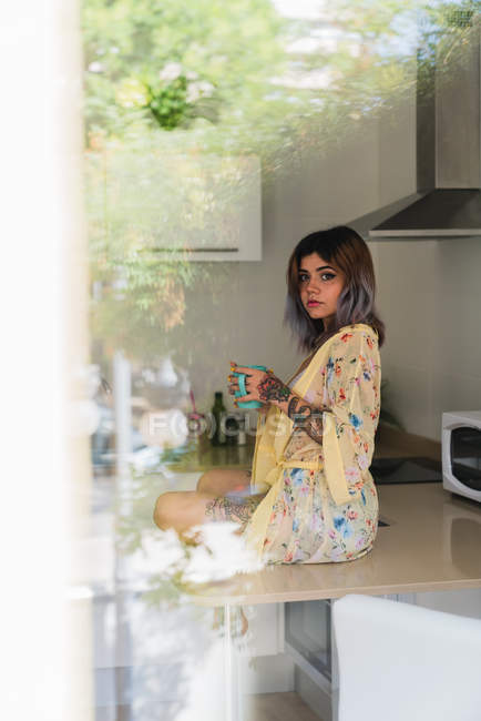 Giovane donna in abito di seta in possesso di tazza di bevanda calda e guardando la fotocamera mentre seduto sul tavolo della cucina dietro il vetro della finestra — Foto stock