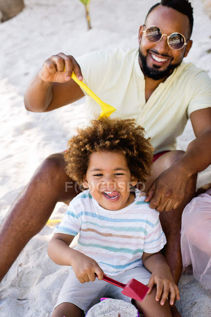 Cheerful adult ethnic man in sunglasses sitting in sand and playing with son on vacation — Stock Photo