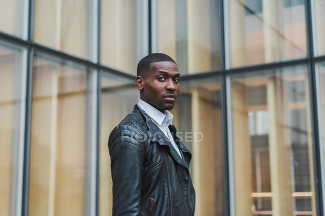 Confident ethnic man standing against glass building and looking at camera — Stock Photo