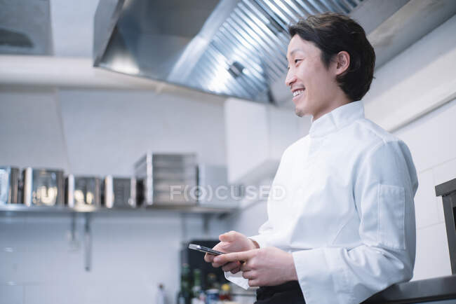 Vista laterale del bell'uomo asiatico in uniforme da chef in piedi in cucina ristorante e smartphone di navigazione — Foto stock