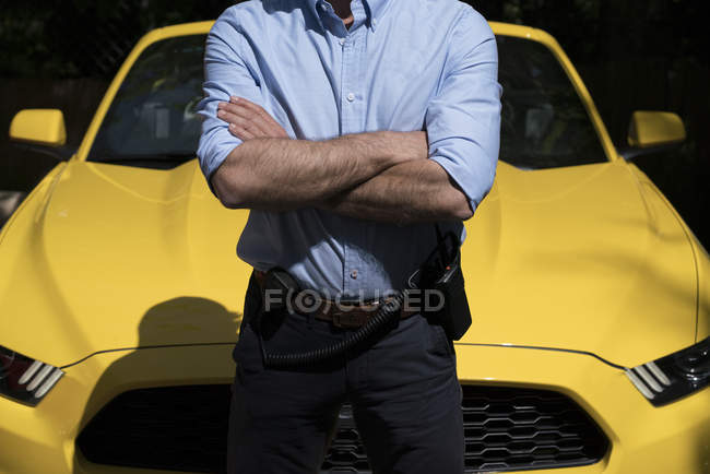 Hombre seguro de sí mismo con radio en el cinturón de pie con los brazos cruzados contra el coche amarillo de lujo moderno - foto de stock