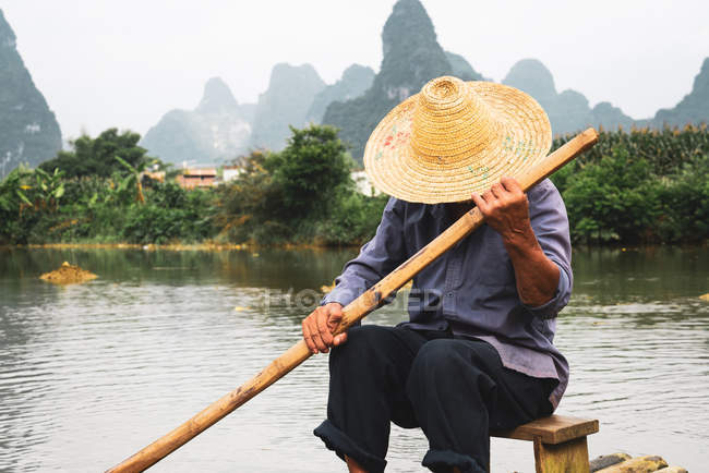 Китайский крестьянин в соломенной шляпе сидит на бамбуковом плоту с горами на заднем плане, Гуанси, Китай — стоковое фото